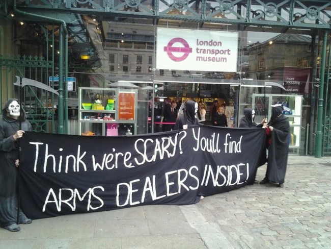 Three protesters dressed as grim reapers outside the London Transport Museum holding a banner saying "Think we're scary? You'll find arms dealers inside!"